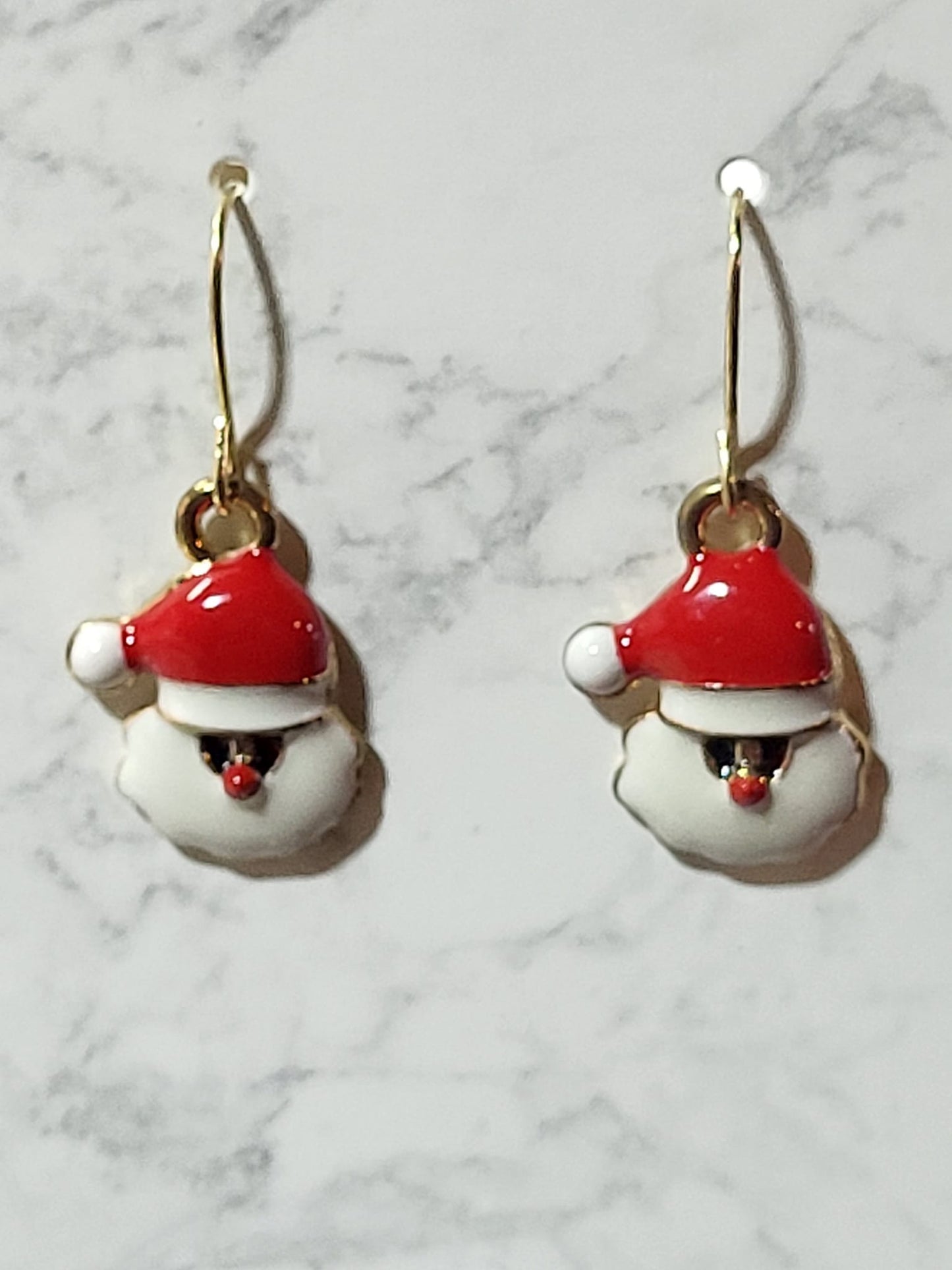 Jolly Santa face enamel charm dangle earrings with glossy red hat, puffy white beard and laughing mouth on gold French wire on marble surface
