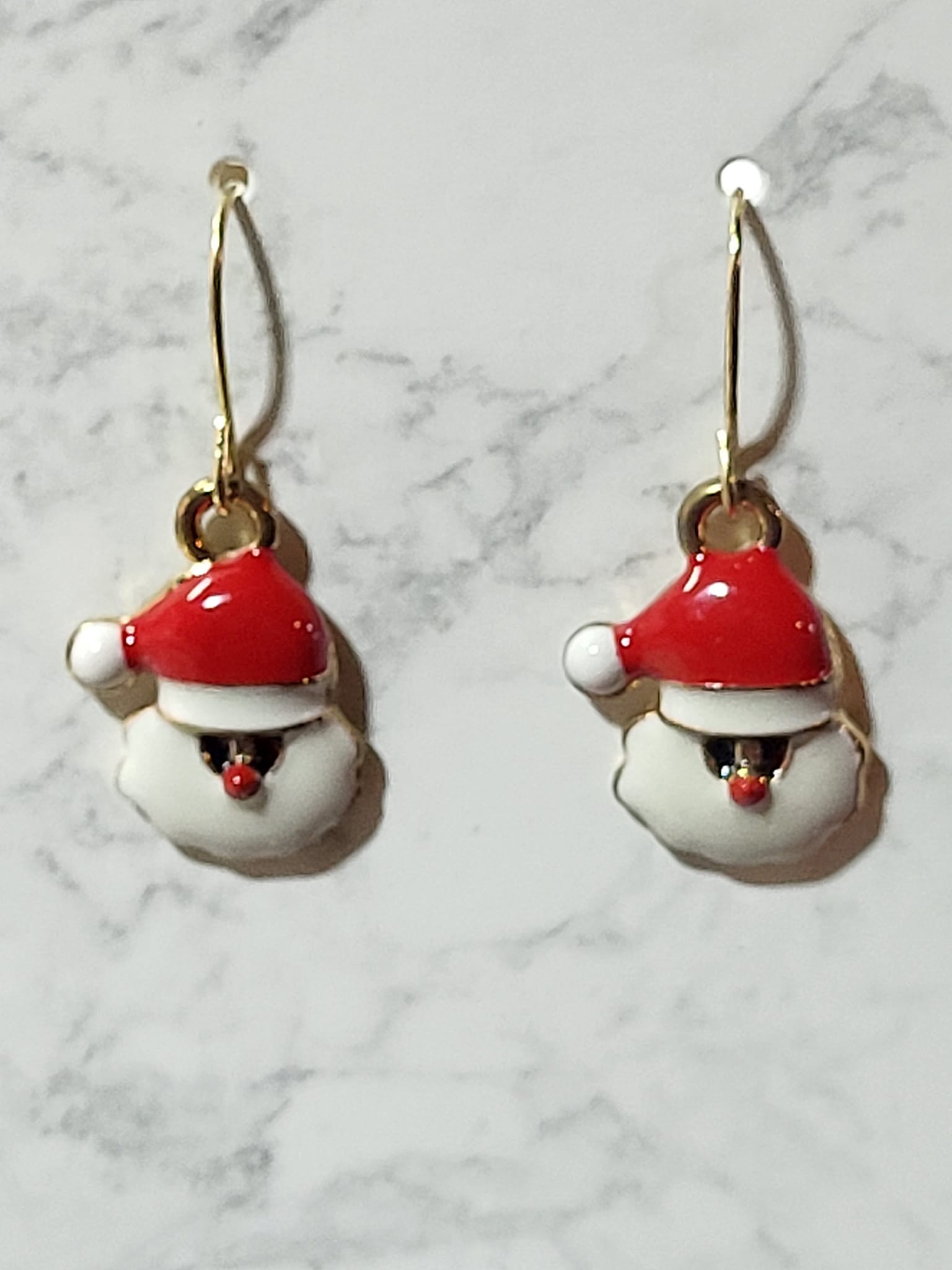 Jolly Santa face enamel charm dangle earrings with glossy red hat, puffy white beard and laughing mouth on gold French wire on marble surface