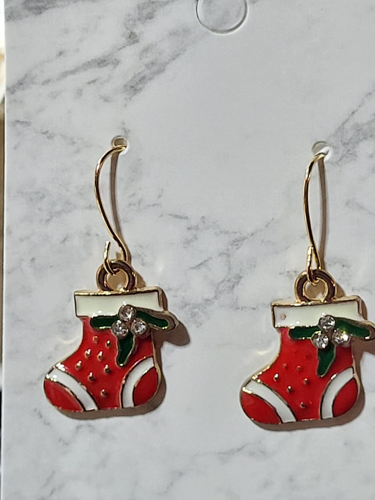 Red enamel Christmas stocking dangle earrings with white cuff, green holly and crystal rhinestone berry accents on gold French wire on marble surface