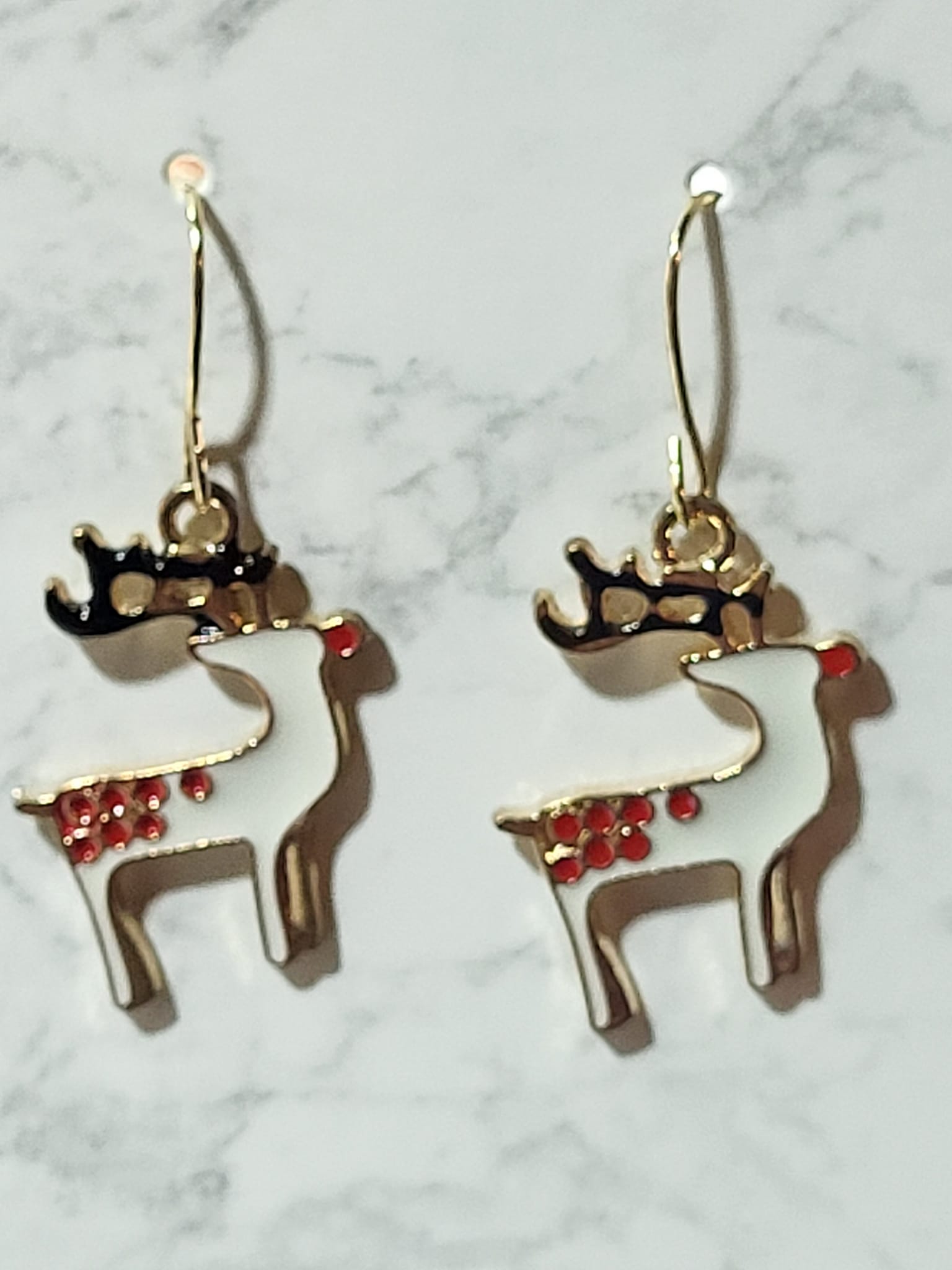 White enamel prancing reindeer dangle earrings with black antlers, red nose and holly berry cluster at neck on gold French wire on marble surface