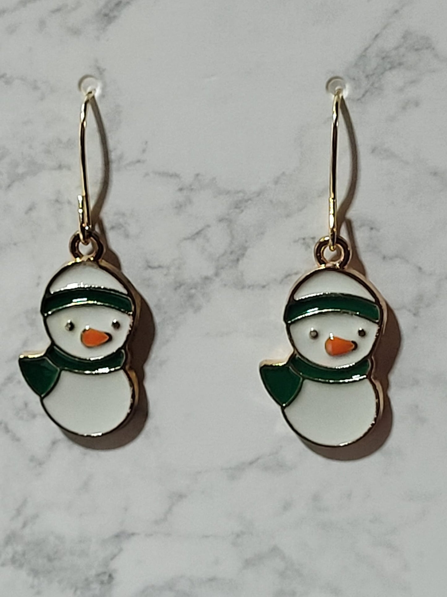 Chubby round snowman bust enamel dangle earrings with dark green beanie hat and scarf and orange carrot nose on gold French wire on marble surface