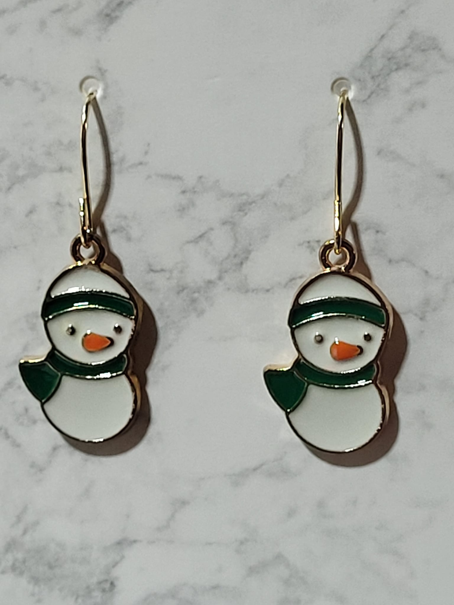 Chubby round snowman bust enamel dangle earrings with dark green beanie hat and scarf and orange carrot nose on gold French wire on marble surface