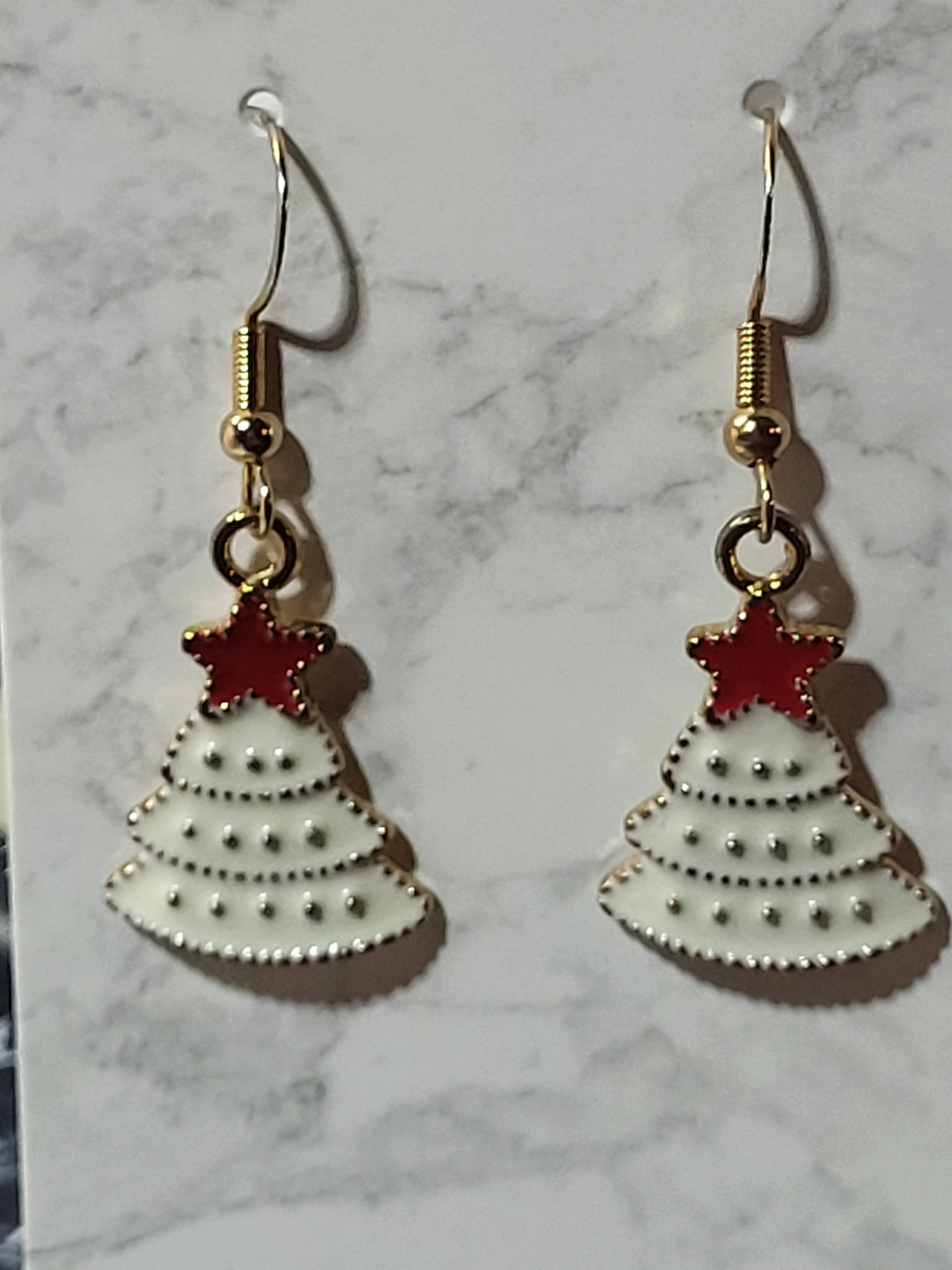 White enamel Christmas tree dangle earrings with gold dot tier detail and red enamel star topper on gold French wire on marble surface