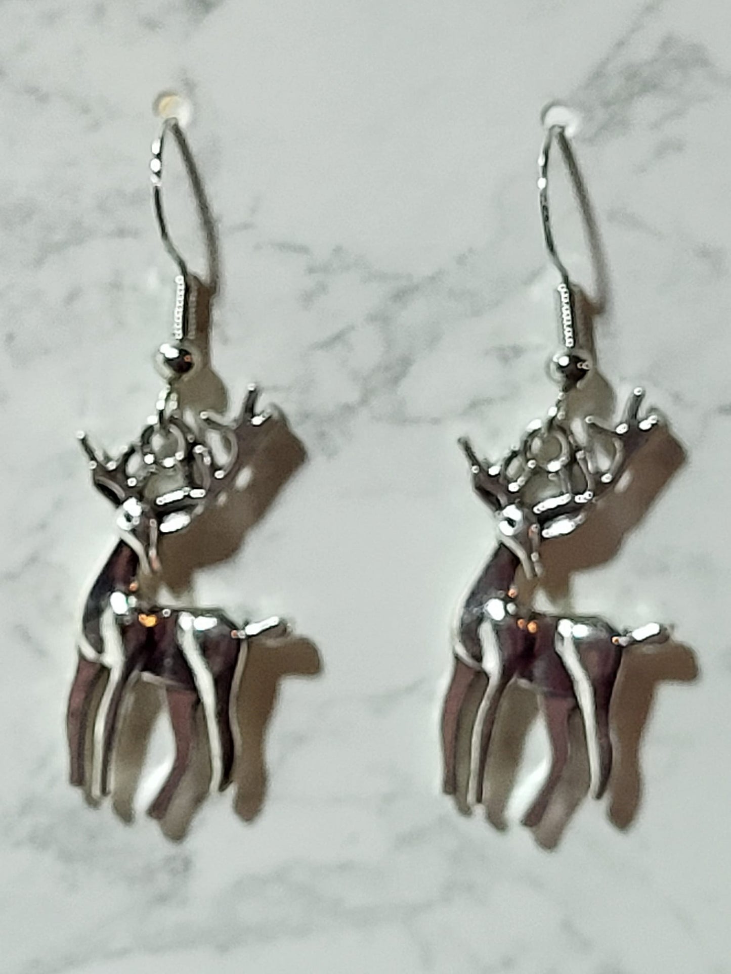 Silver tone full-body reindeer stag charm dangle earrings with detailed antlers and dark enamel body on silver French wire on marble surface