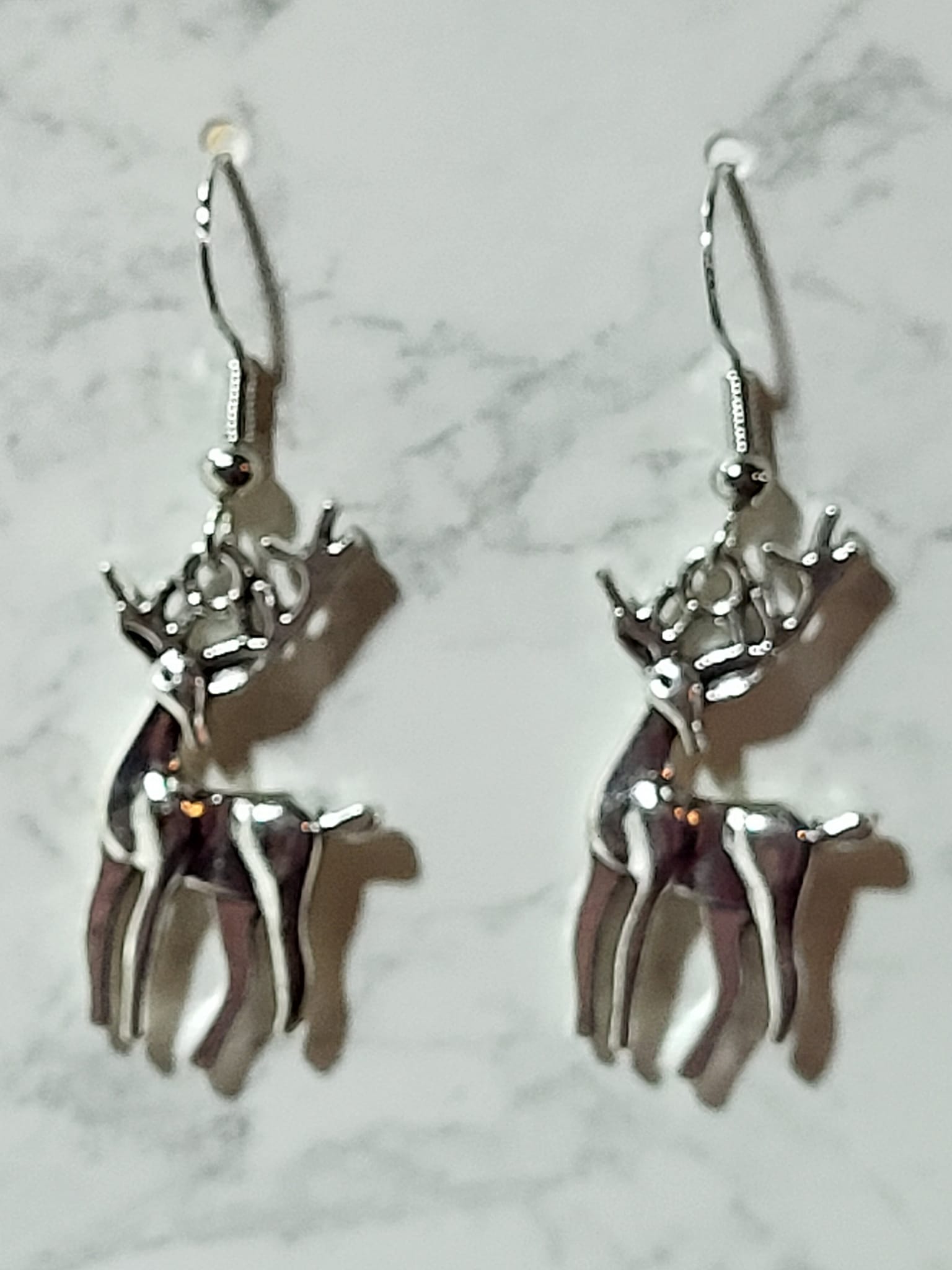 Silver tone full-body reindeer stag charm dangle earrings with detailed antlers and dark enamel body on silver French wire on marble surface