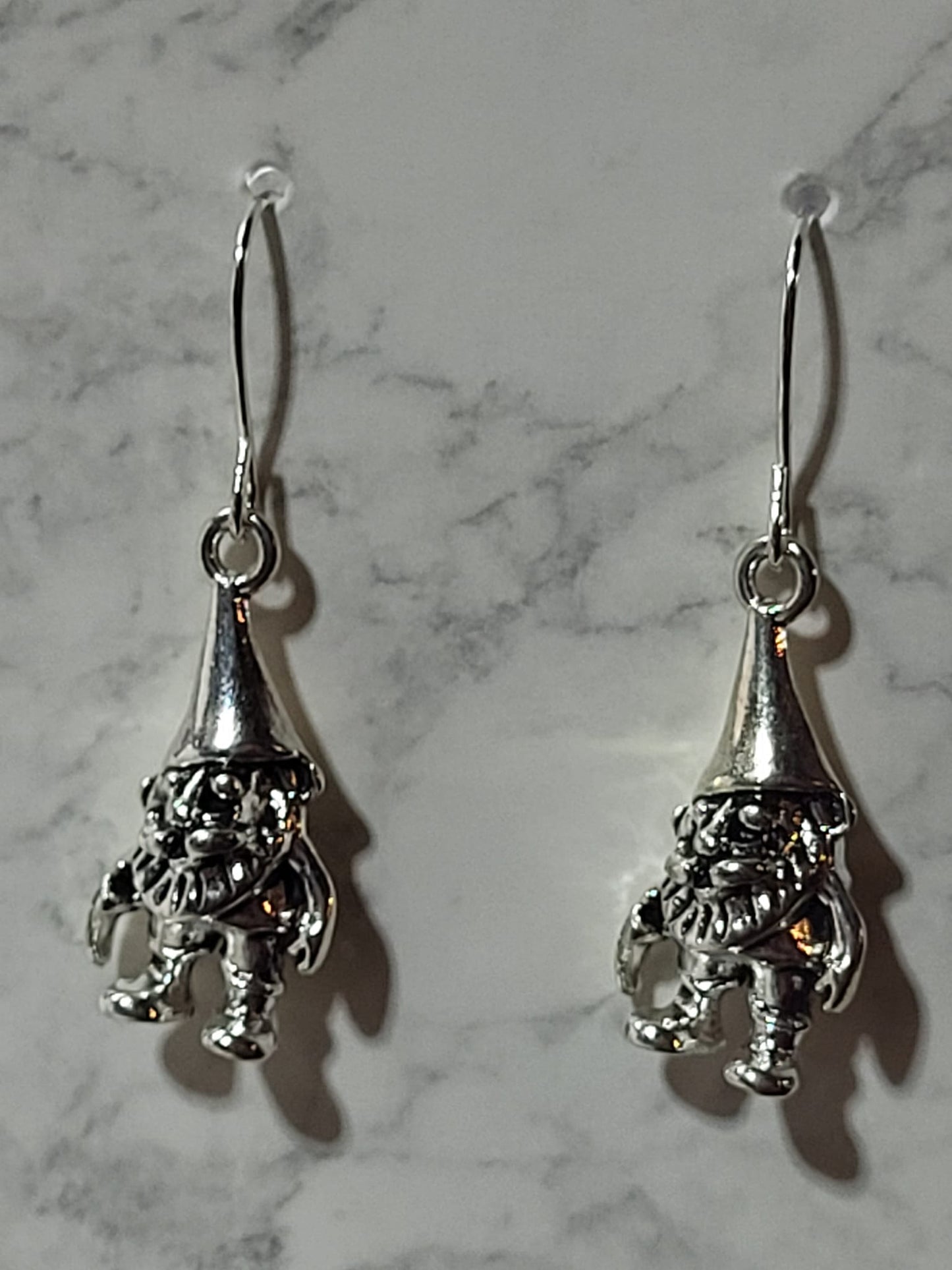 Silver Christmas elf charm dangle earrings with tall pointed hat and detailed boots on silver French wire on marble surface