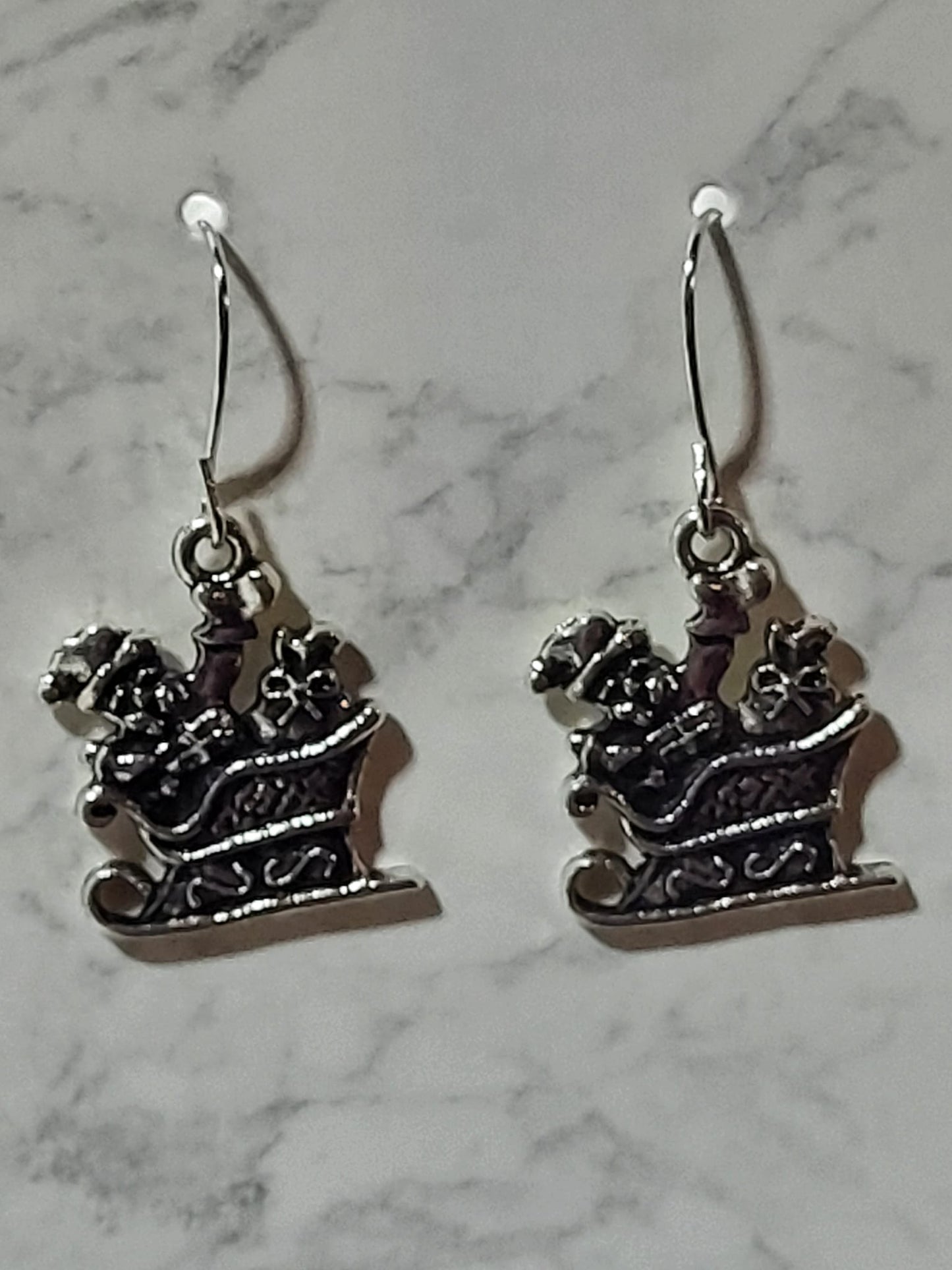 Silver Santa sleigh charm dangle earrings with gift boxes and scrollwork detail on silver French wire on marble surface