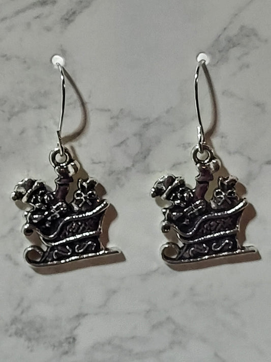 Silver Santa sleigh charm dangle earrings with gift boxes and scrollwork detail on silver French wire on marble surface
