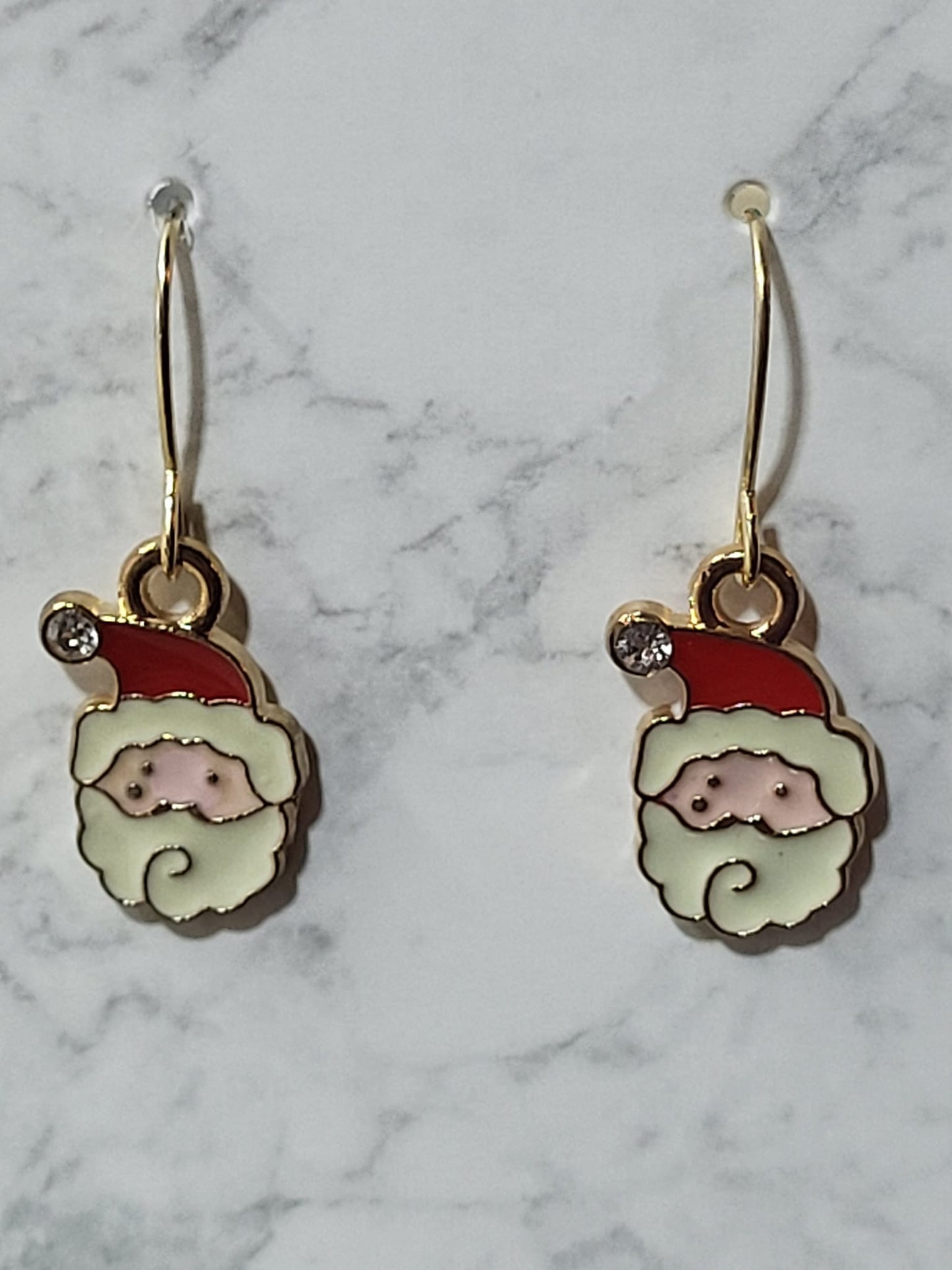 Enamel Santa face charm dangle earrings with red hat, white beard, rosy nose and crystal accent on gold French wire on marble surface