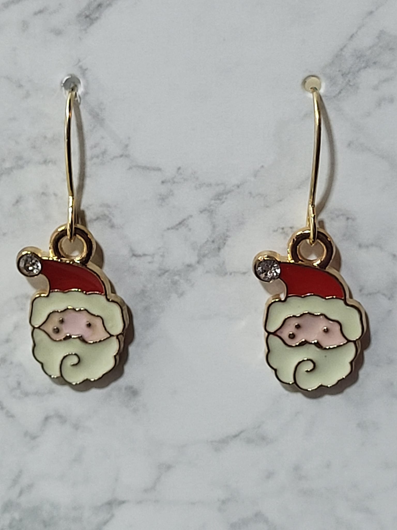 Enamel Santa face charm dangle earrings with red hat, white beard, rosy nose and crystal accent on gold French wire on marble surface