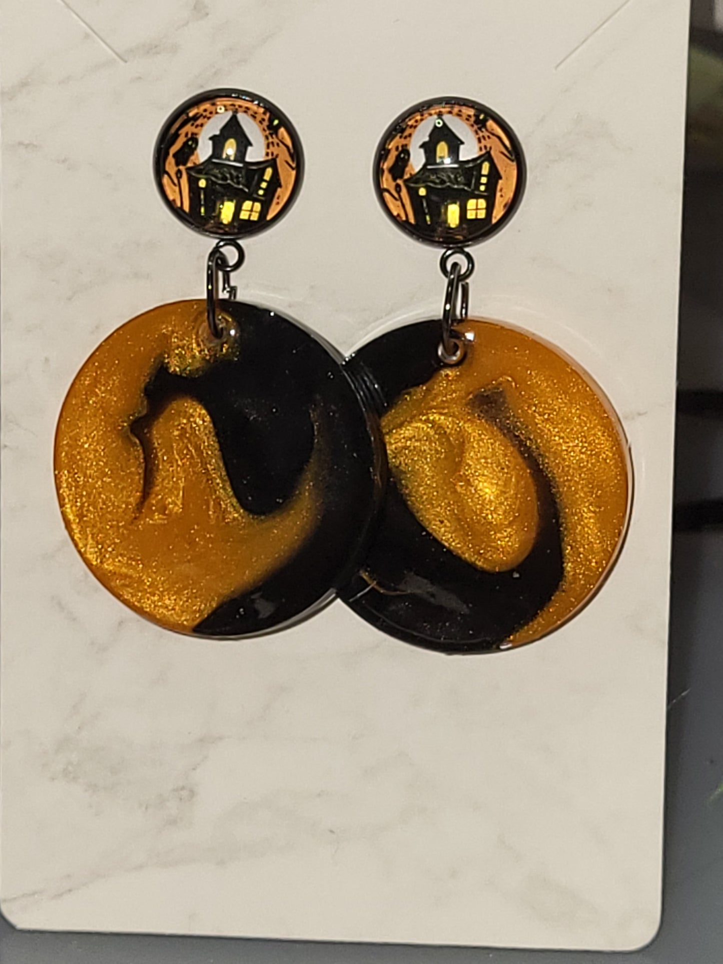 Black and gold glitter swirl resin circle dangle earrings with orange haunted house photo cab studs on marble display card