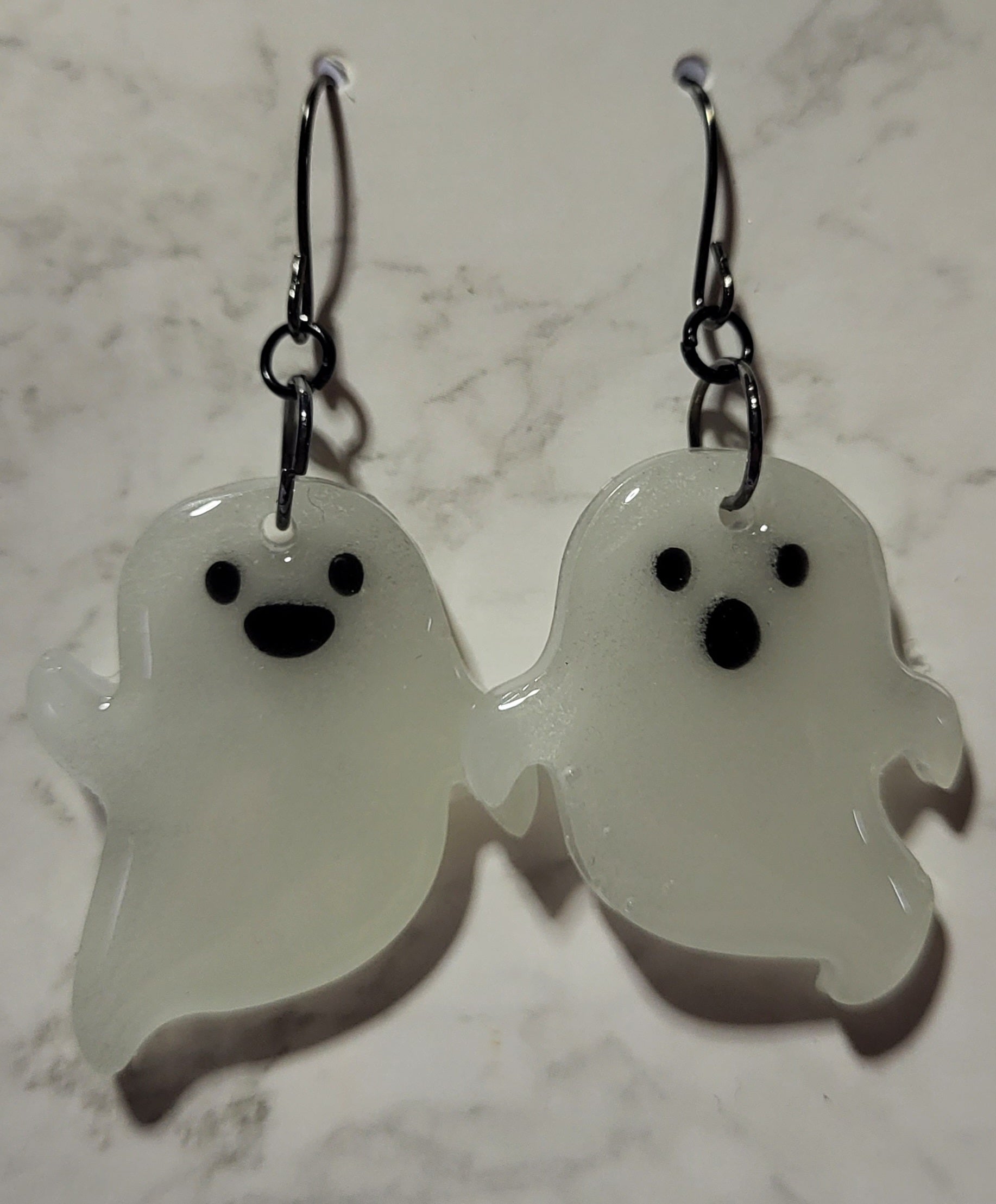Chunky mismatched ghost glow in the dark resin earrings in daylight showing bright white resin with black facial features on marble surface