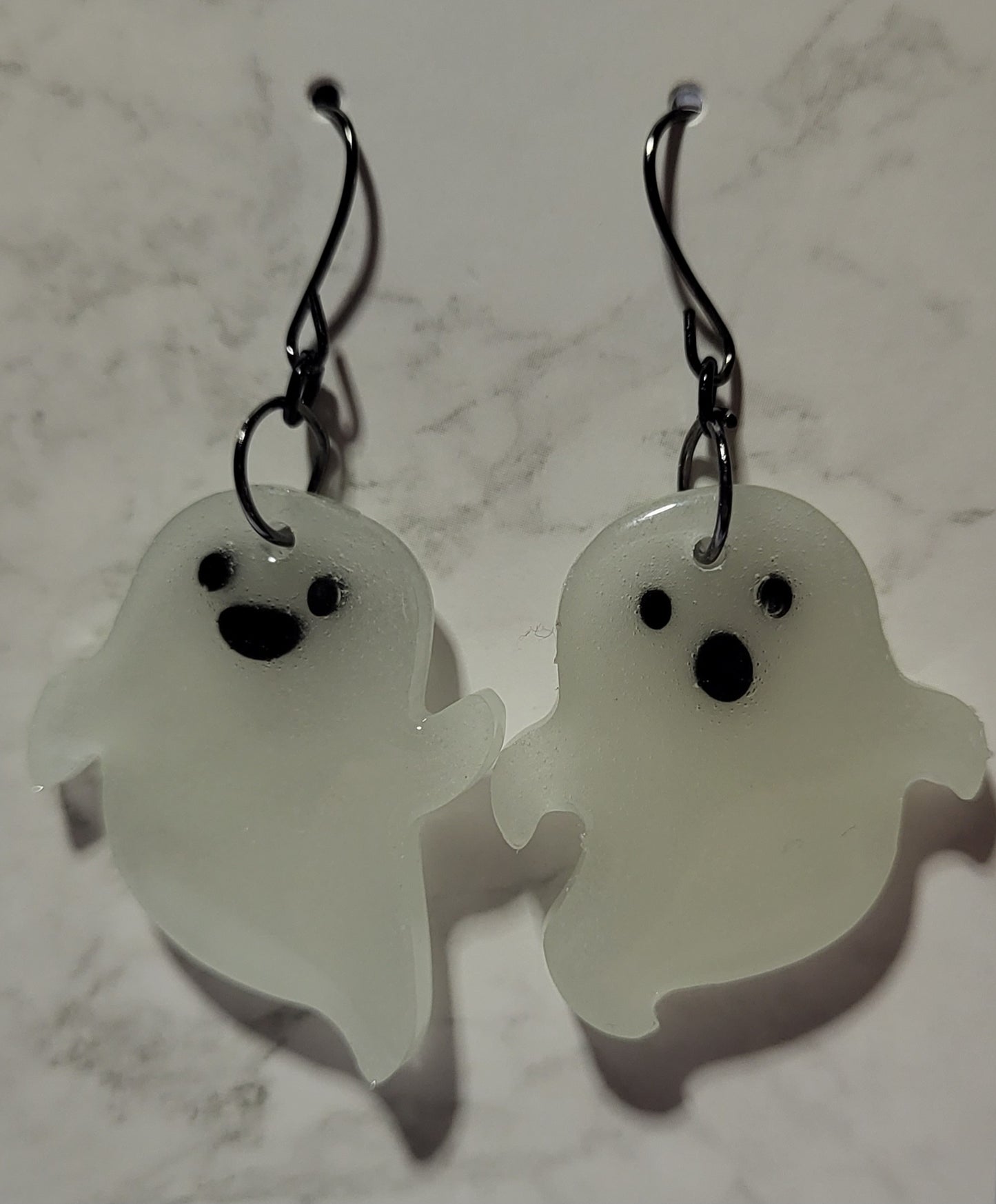 Mismatched ghost glow in the dark resin earrings in daylight showing translucent white with black facial features on marble surface