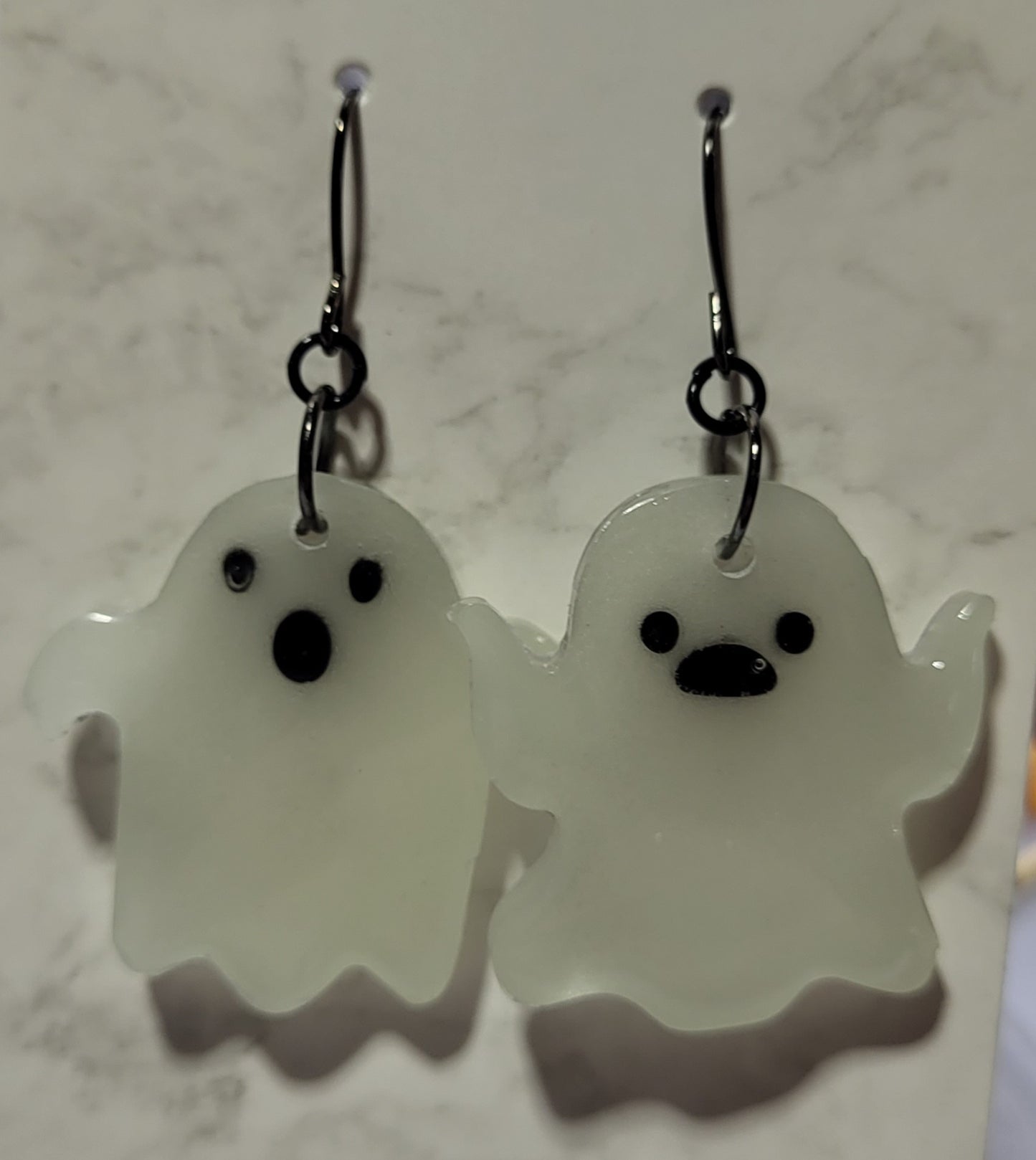Chunky mismatched ghost glow in the dark resin earrings in daylight showing bright white resin with black facial features on marble surface