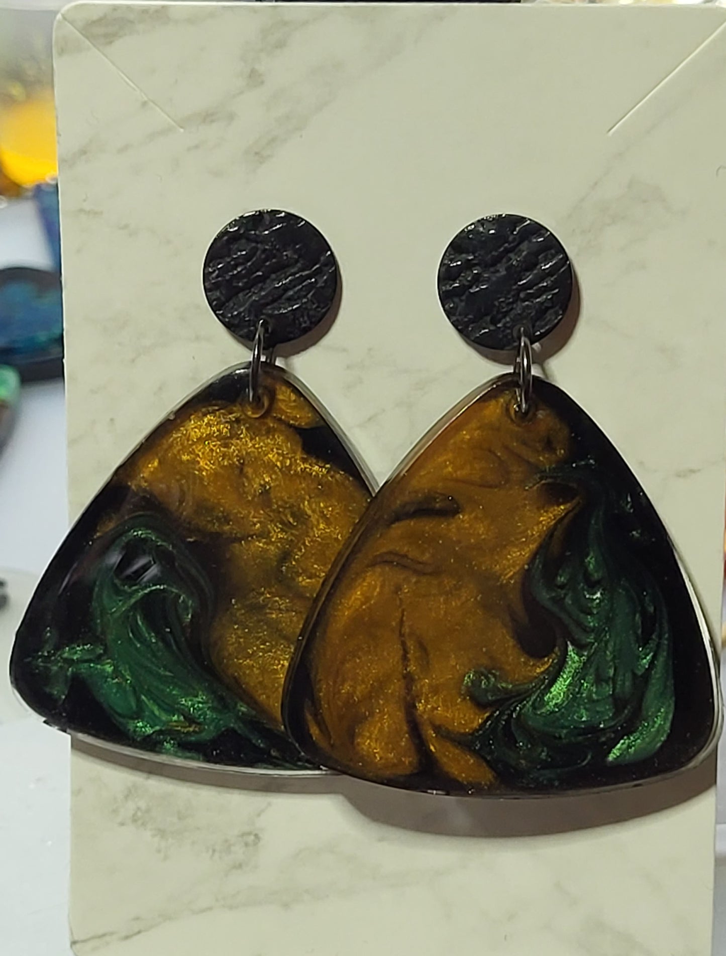 Witches brew triangle resin drop earrings on marble card showing amber gold and emerald green swirl on black base with black textured circle stud top