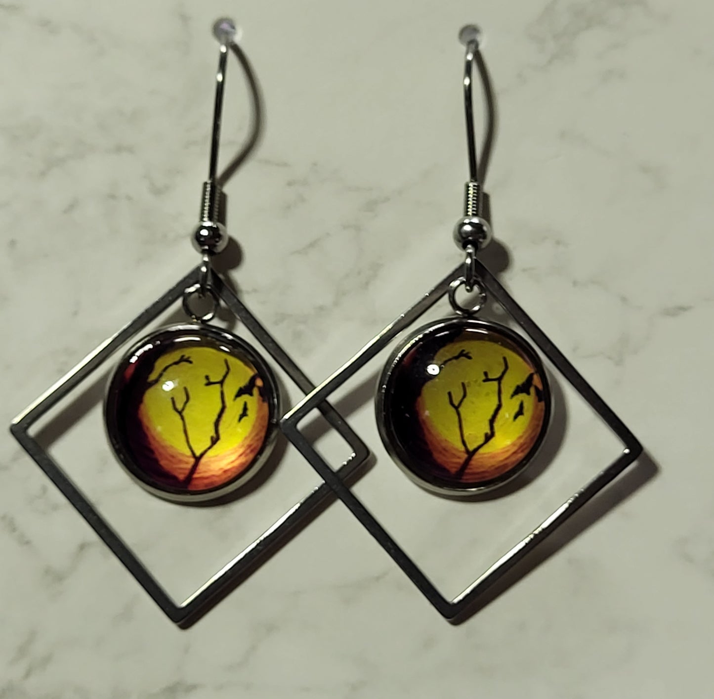 Halloween night sky glass dome cab dangle earrings in open silver diamond frame on marble surface showing glowing orange sky with silhouetted bats and bare trees