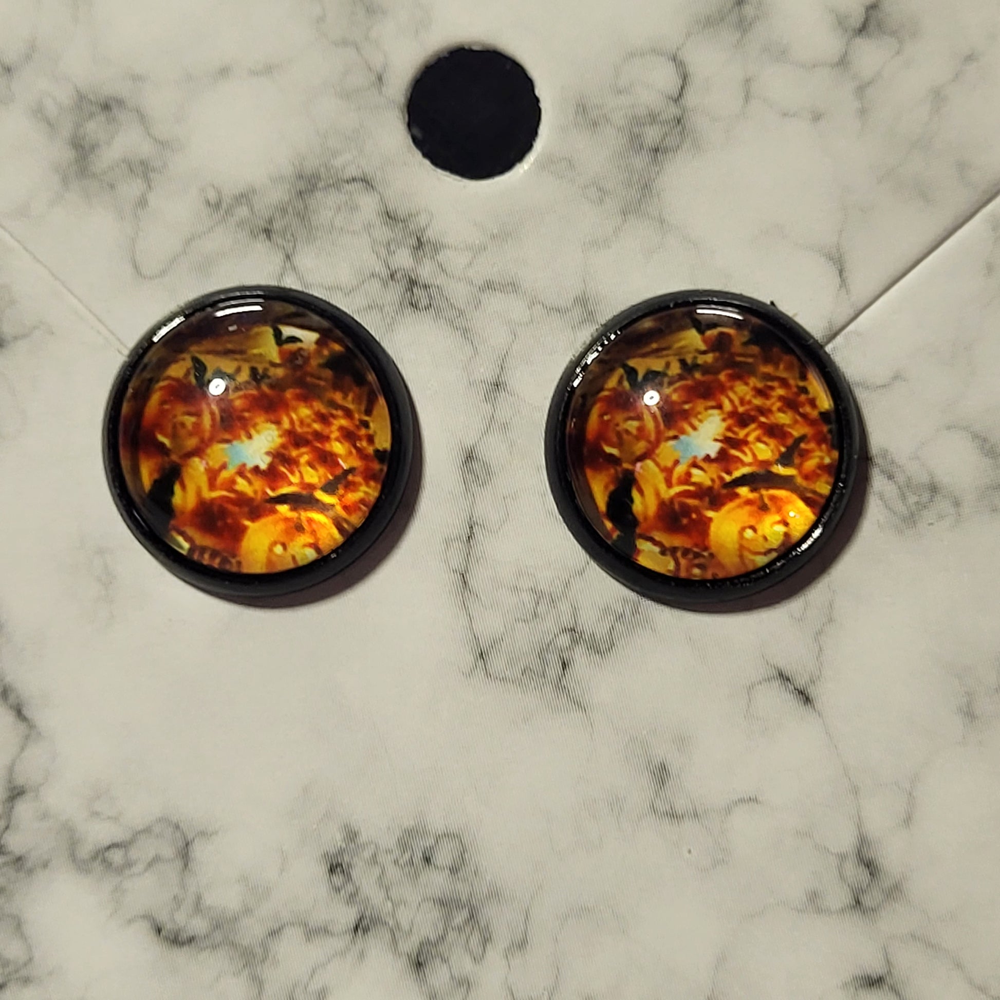 Round glass dome jack-o-lantern pumpkin stud earrings in black bezel on marble card showing vibrant Halloween scene with bats and autumn leaves