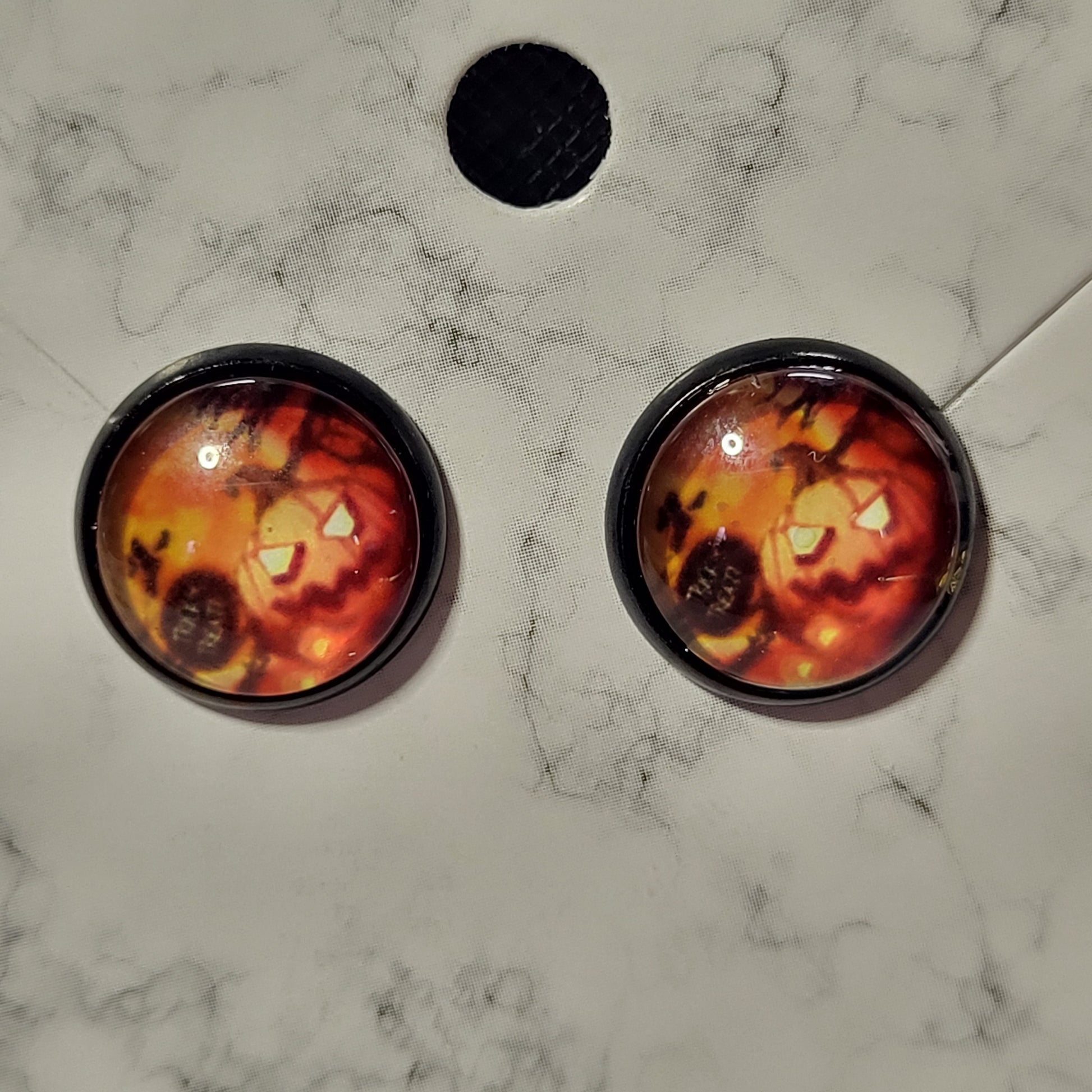 Round glass dome stud earrings in black bezel featuring close-up glowing jack-o-lantern face with triangle eyes and jagged mouth in deep red and amber tones