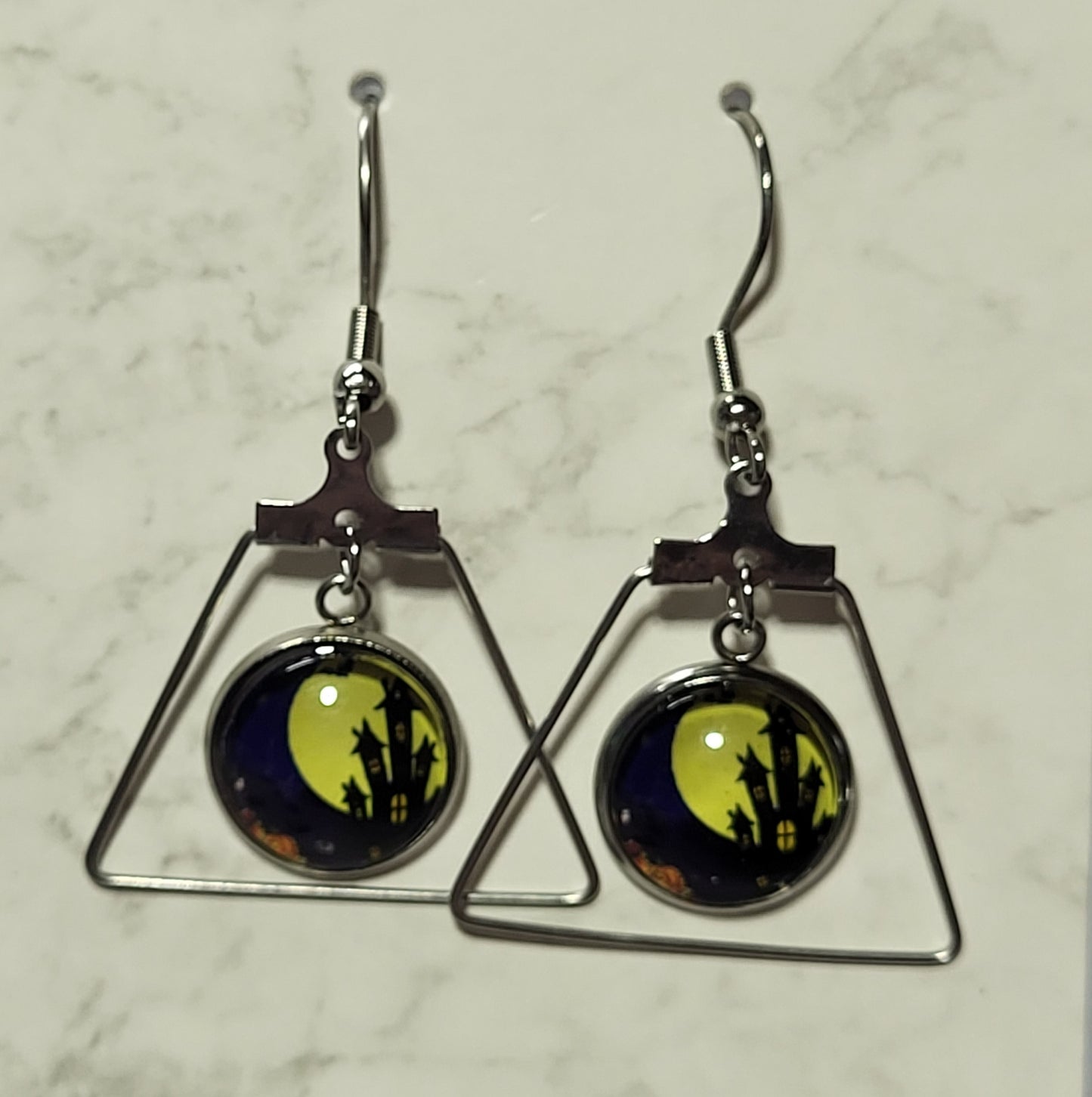 Full moon haunted castle glass dome cab earrings suspended inside open silver trapezoid triangle frame on French wire hook on marble surface