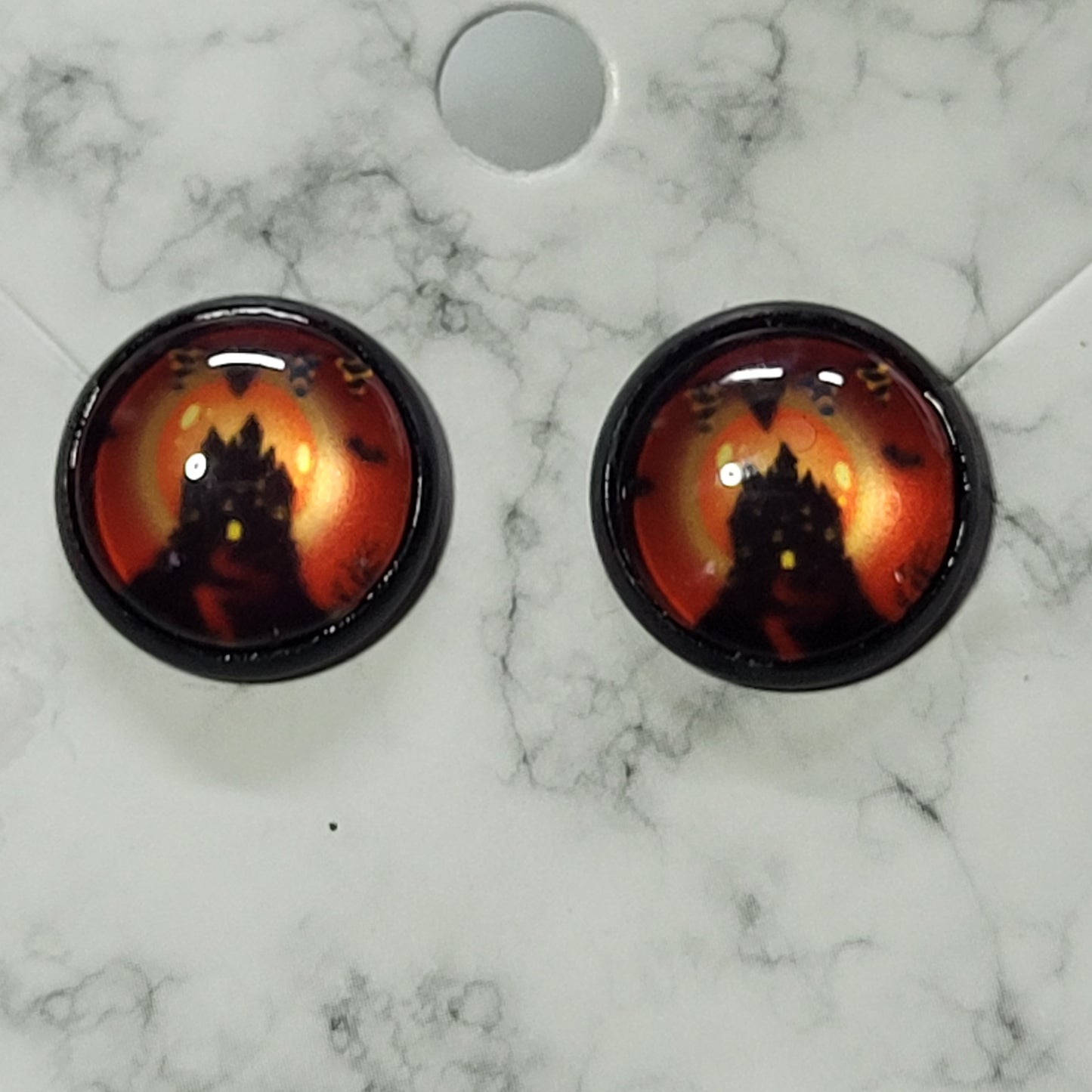 Round glass dome stud earrings in black bezel featuring silhouetted haunted castle against deep fiery orange-red Halloween night sky with bats