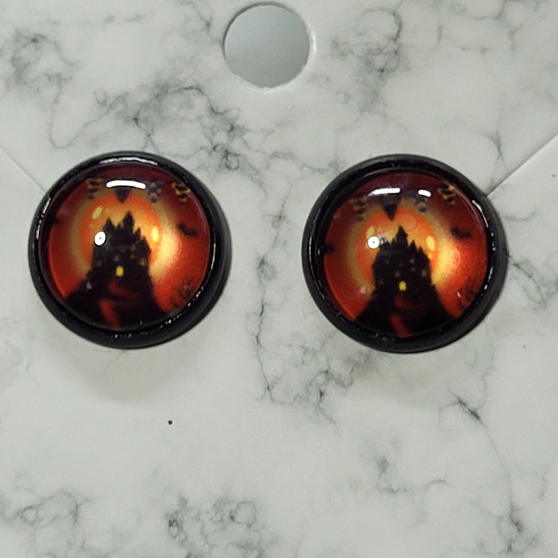 Round glass dome stud earrings in black bezel featuring silhouetted haunted castle against deep fiery orange-red Halloween night sky with bats