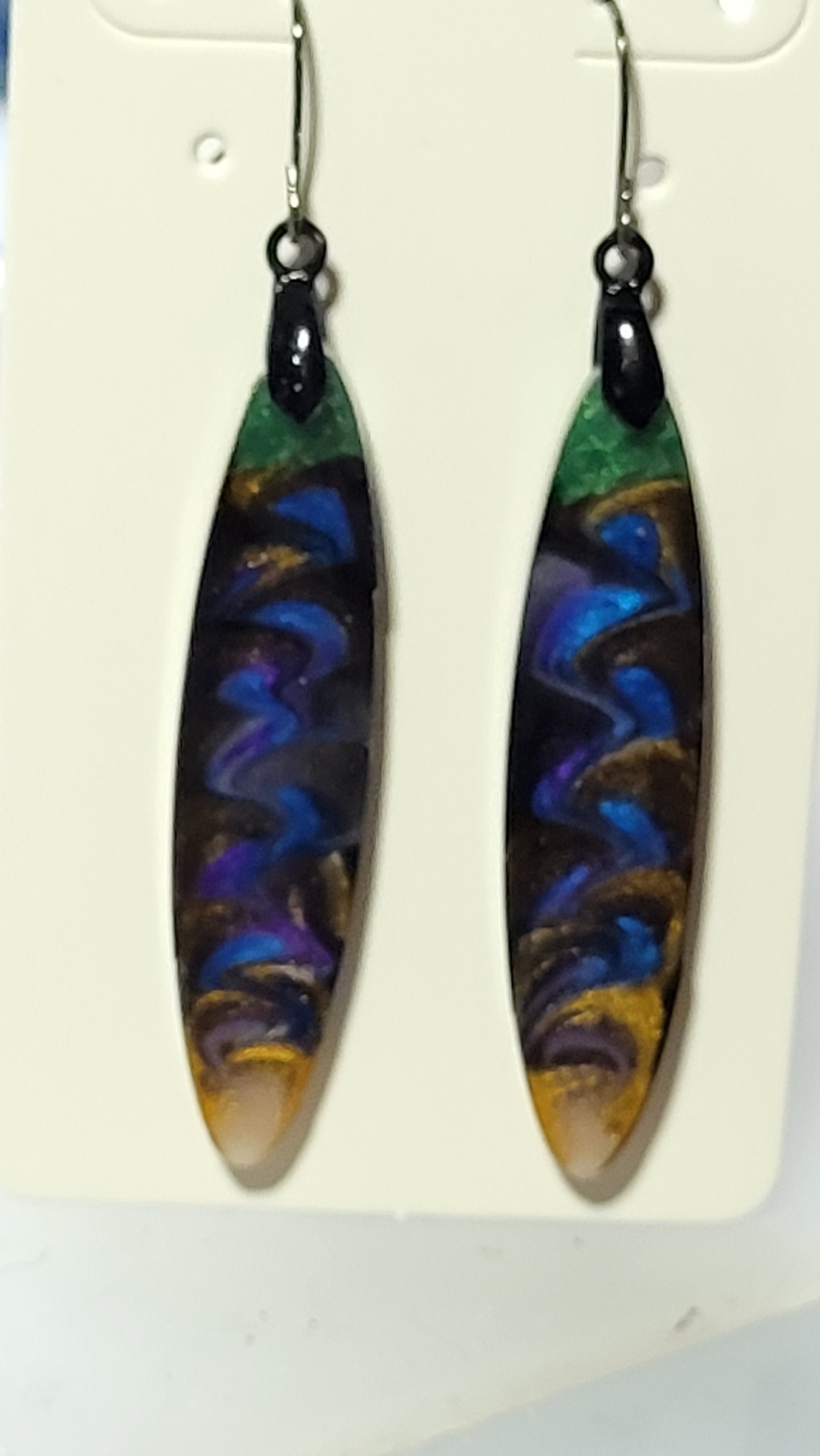 Long slim surfboard resin dangle earrings with electric blue and purple zigzag wave pattern, emerald green top and amber gold tip on white earring card