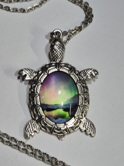 Silver-tone turtle necklace with green and purple northern lights imagery on a white surface.