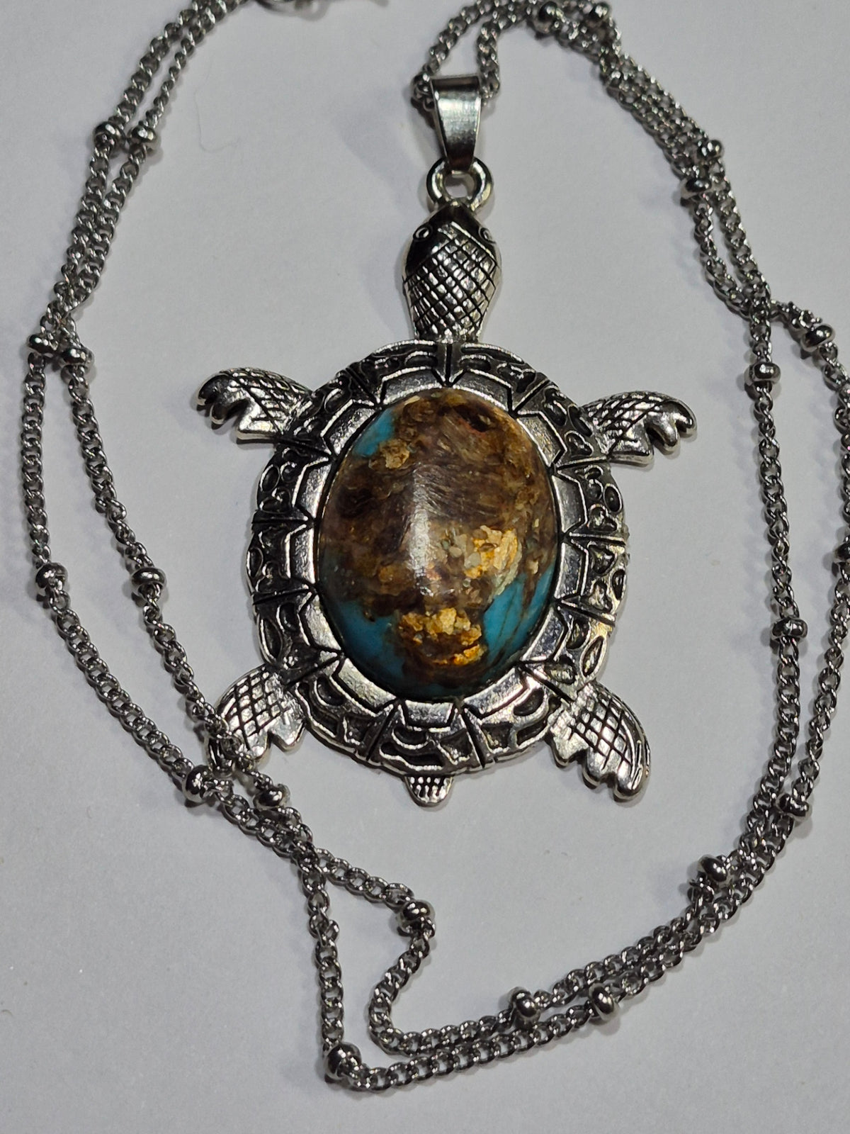 Turquoise-blue and brown sea turtle pendant surrounded by its silver-tone chain on white.