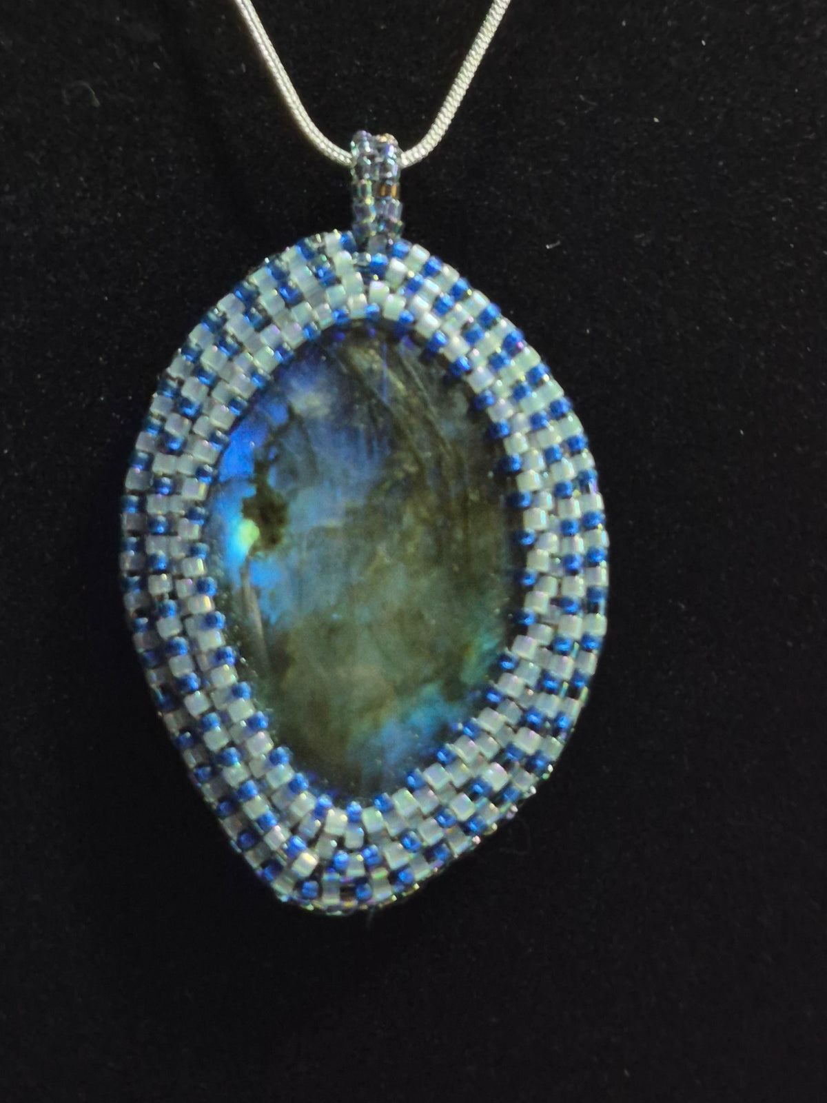 Oval blue-flash cabochon pendant with a blue and white beadwoven bezel on a black background.
