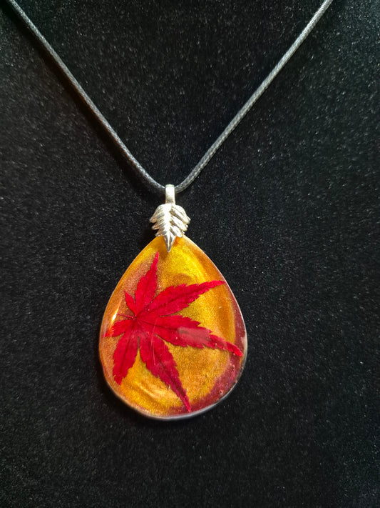 Japanese maple leaf resin teardrop pendant with gold shimmer hanging from a black cord against black.