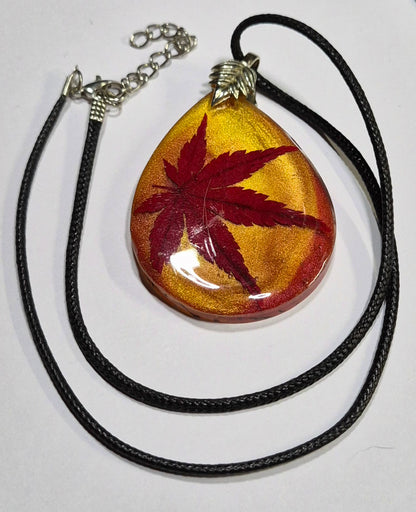 Maple leaf resin pendant with black faux leather cord and extender laid out on white.