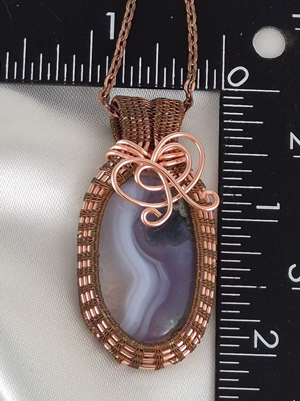 Banded oval stone pendant with woven copper frame beside a ruler for scale.