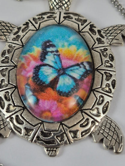 Close-up of a blue butterfly over colorful flowers in the cabochon shell of a silver-tone turtle pendant.