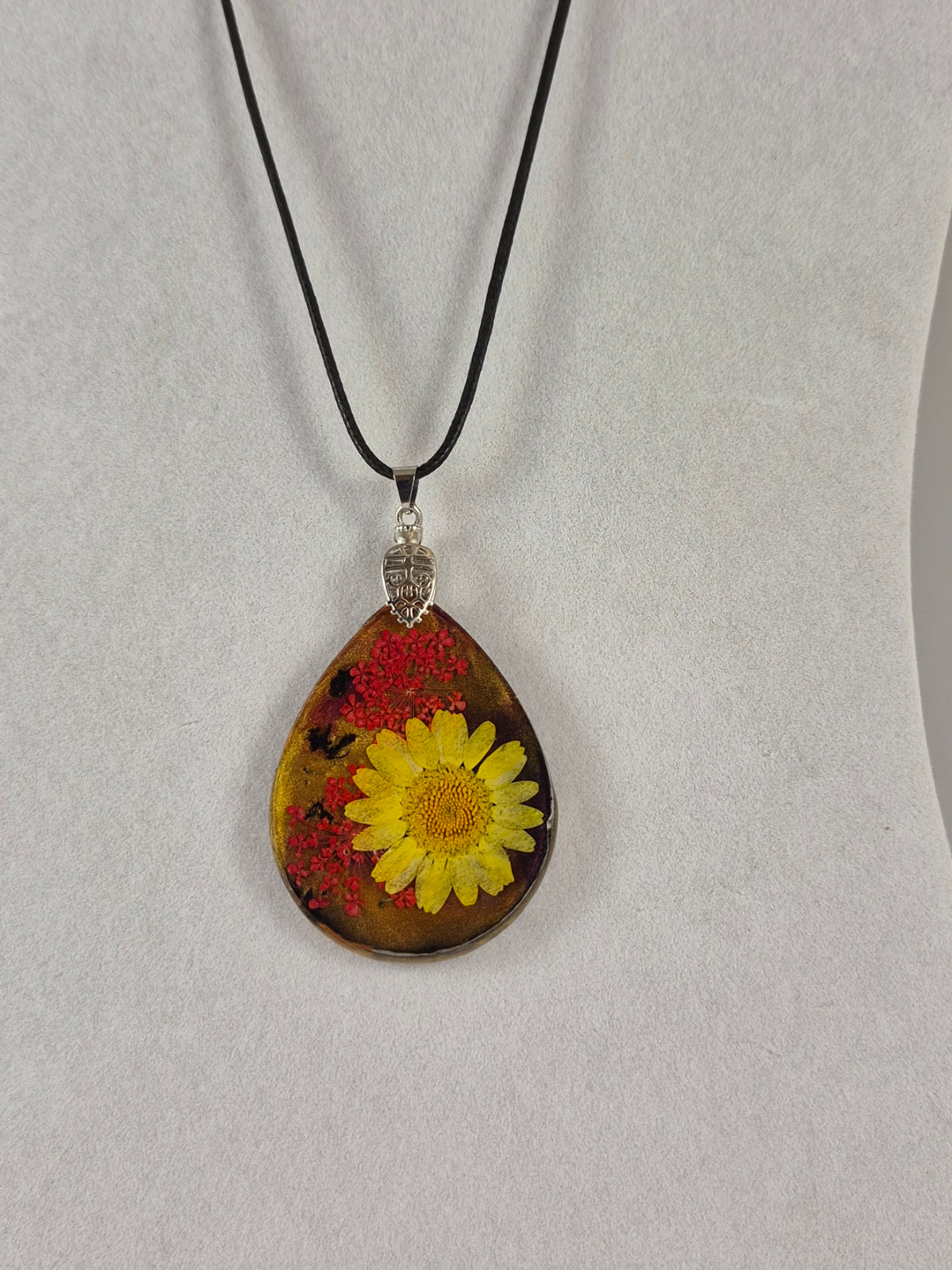 Teardrop resin pendant with a yellow flower and red botanical accents hanging on a black cord.