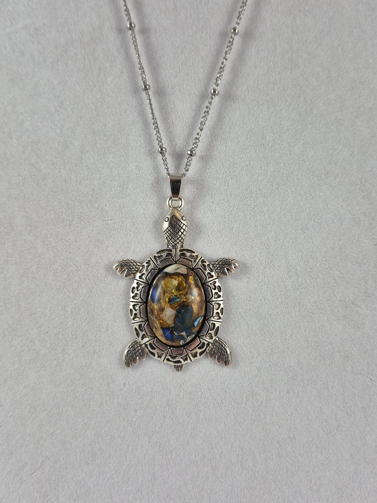 Silver-tone sea turtle pendant with a blue, cream, and gold-toned mosaic-style cabochon hanging on a chain.