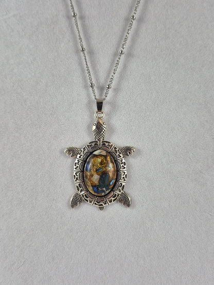 Silver-tone sea turtle pendant with a blue, cream, and gold-toned mosaic-style cabochon hanging on a chain.